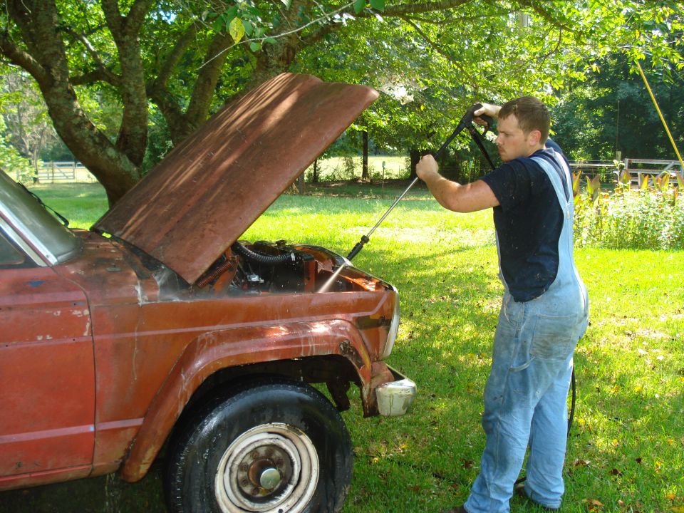 Man pressure washing the engine bay of a rusty 1984 Jeep J-10 with the hood open