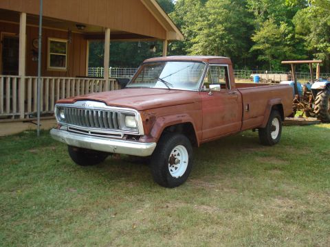 1984 Jeep J-10 - A vintage Jeep J-10 pickup truck sits on a grassy lawn next to a house, with a tractor visible in the background.