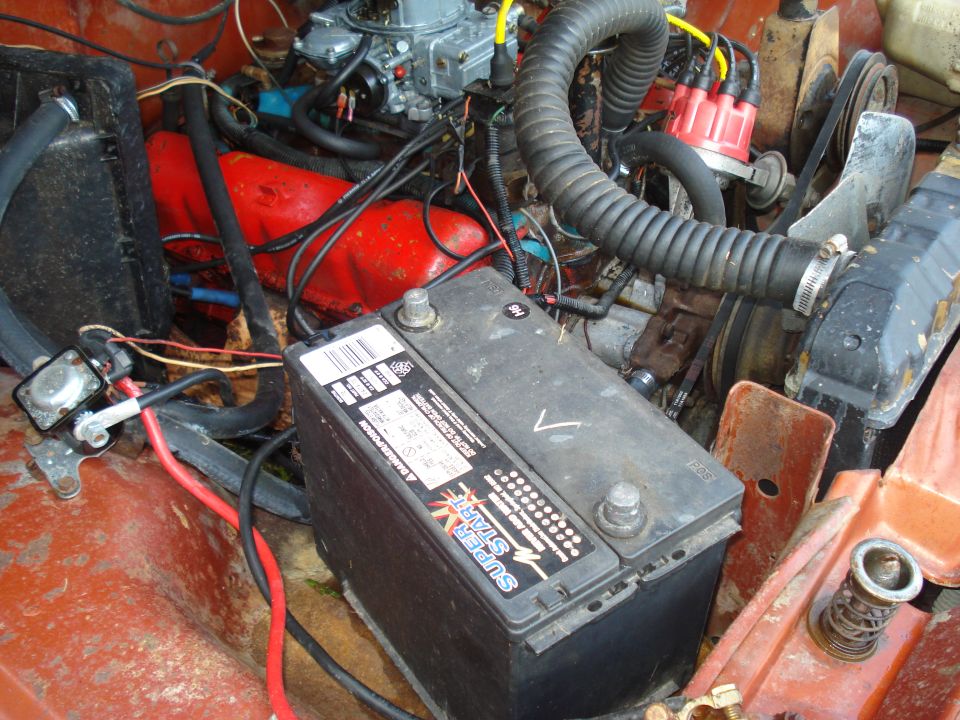 Engine bay with a black battery, red engine block, carburetor, and hoses. Rusty metal surfaces are visible