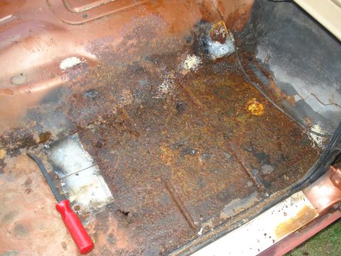 1984 Jeep J-10 - The floor pan of a vehicle shows extensive rust damage and a red-handled tool resting on a newly patched section.