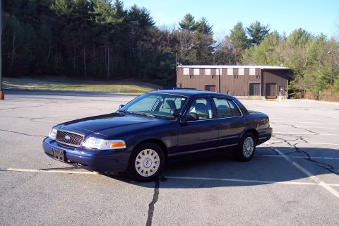 2001 Ford Crown Victoria - A dark blue 2001 Ford Crown Victoria is parked in a paved lot with a building and trees in the background.