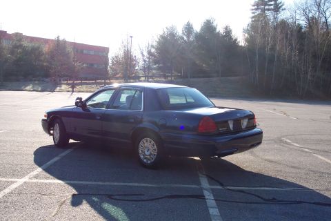 2001 Ford Crown Victoria - A dark blue sedan is parked in an empty lot with a building and trees in the background.
