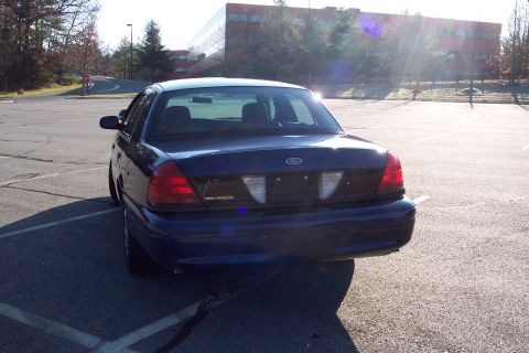 2001 Ford Crown Victoria - The rear of a dark blue 2001 Ford Crown Victoria sedan is visible in a parking lot with a modern building in the background.
