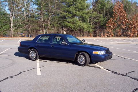 2001 Ford Crown Victoria - A dark blue sedan is parked in an empty lot with trees in the background.