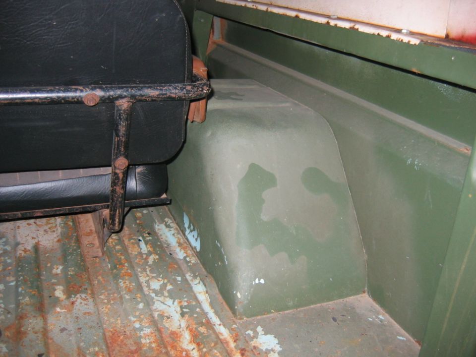 Rear interior of a vehicle showing a black seat, green painted wheel well, and rusted floor