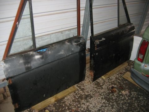 1992 Ford Mustang - Two removed black early Ford Bronco doors leaning against a corrugated metal wall inside a carport, resting on treated lumber over gravel.…