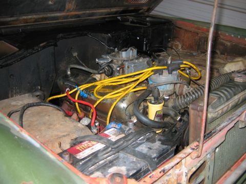 1992 Ford Mustang - Grimy engine bay of a green vintage 4x4 with the hood up, showing an inline six with a two-barrel carburetor, bright yellow plug wires, a y…