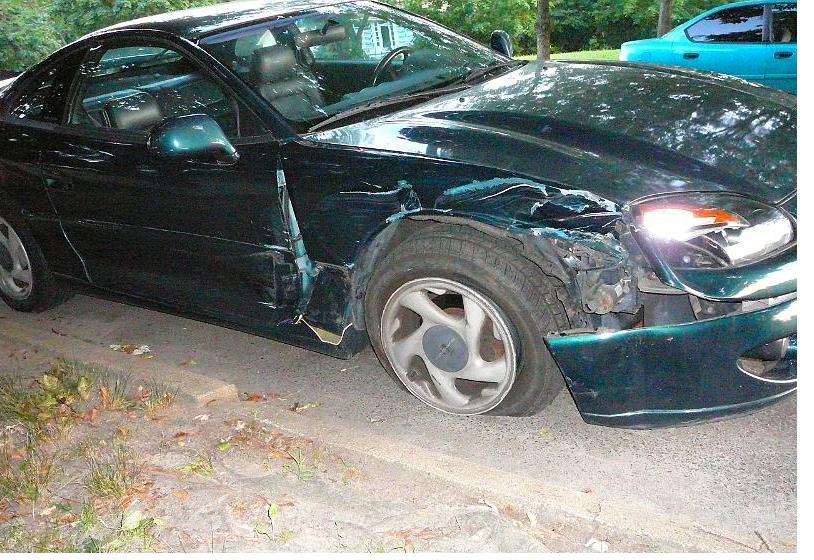 Damaged green coupe, front fender and door crumpled. Flat tire. Another blue car visible in background