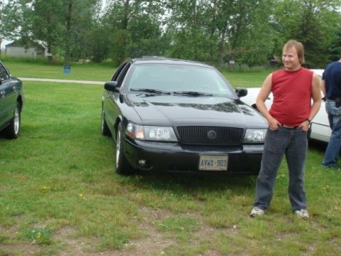 2004 Ford Crown Victoria - A man stands beside a black Mercury Grand Marquis parked on a grassy area with trees in the background.