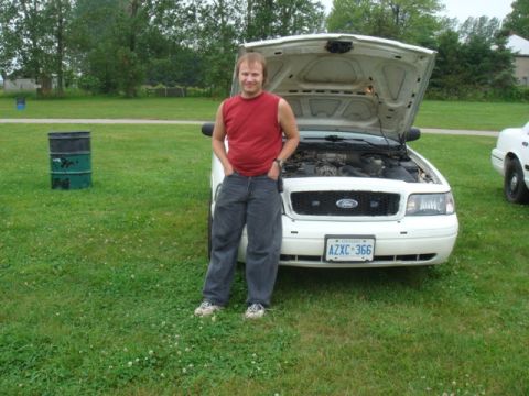 2004 Ford Crown Victoria - A man stands next to a white 2004 Ford Crown Victoria with its hood up, showing the engine compartment.