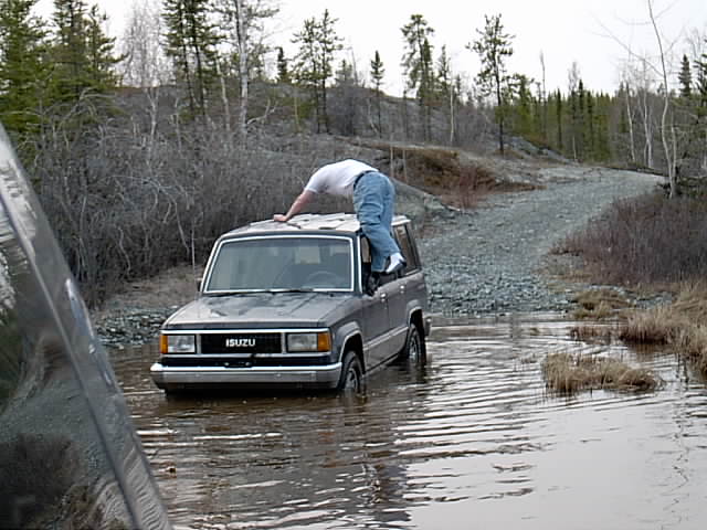 Silver Isuzu Trooper stuck in a flooded gravel trail as a person climbs out the driver's window