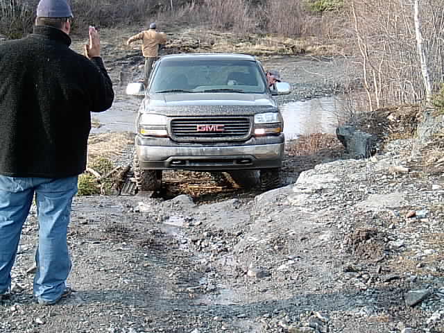 Muddy pewter GMC Sierra pickup exiting a water crossing with a spotter guiding