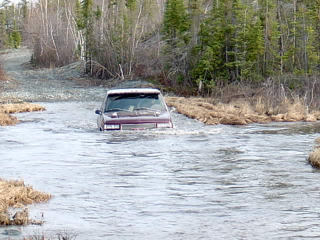 Maroon vehicle fording deep flooded stream, water near windows, on wooded gravel road