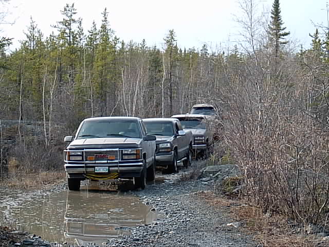 White GMC Sierra pickup with grille guard leading a line of trucks on a muddy gravel bush road