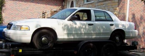 2003 Ford Crown Victoria - This white sedan is being transported on a trailer, with a brick house in the background.