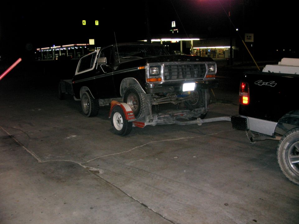 Black 1978 Ford Bronco on a trailer, towed by a black pickup truck at night