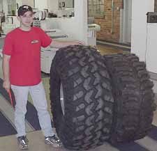 1990 Ford Bronco - A man stands next to two large off-road tires, one of which has the SuperMotors logo visible.