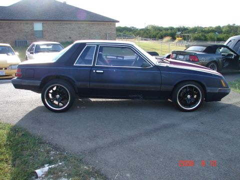1985 Ford Mustang - Side view of a dark blue coupe, possibly a 1985 Ford Mustang, with aftermarket wheels.
