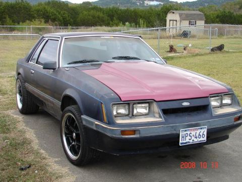 1985 Ford Mustang - A 1985 Ford Mustang coupe is parked outside on a paved surface with a grassy area and a fence in the background.