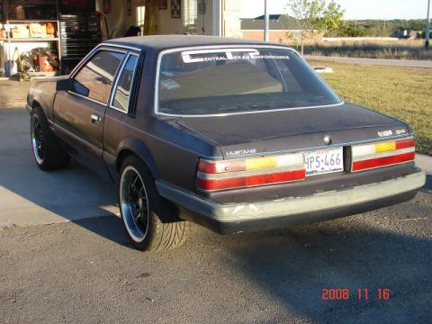 1985 Ford Mustang - A dark coupe, identified as a 1985 Ford Mustang, is parked outside a garage, showing its rear-left side and side profile.
