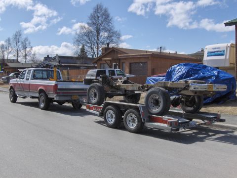 1990 Ford Bronco - Ford pickup tows a trailer carrying a vehicle chassis and a tarp-covered vehicle.