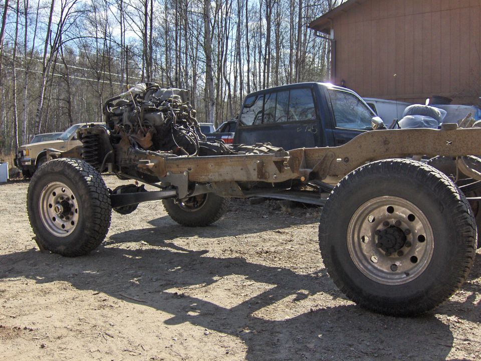 Bodyless 4x4 rolling chassis with V8 and mud tires parked on gravel outside a wood building
