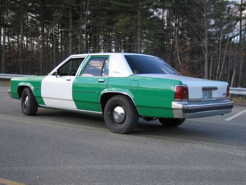 1991 Ford LTD Crown Victoria - Green-and-white two-tone Ford sedan seen from the rear quarter, parked on a road with a guardrail and bare trees.