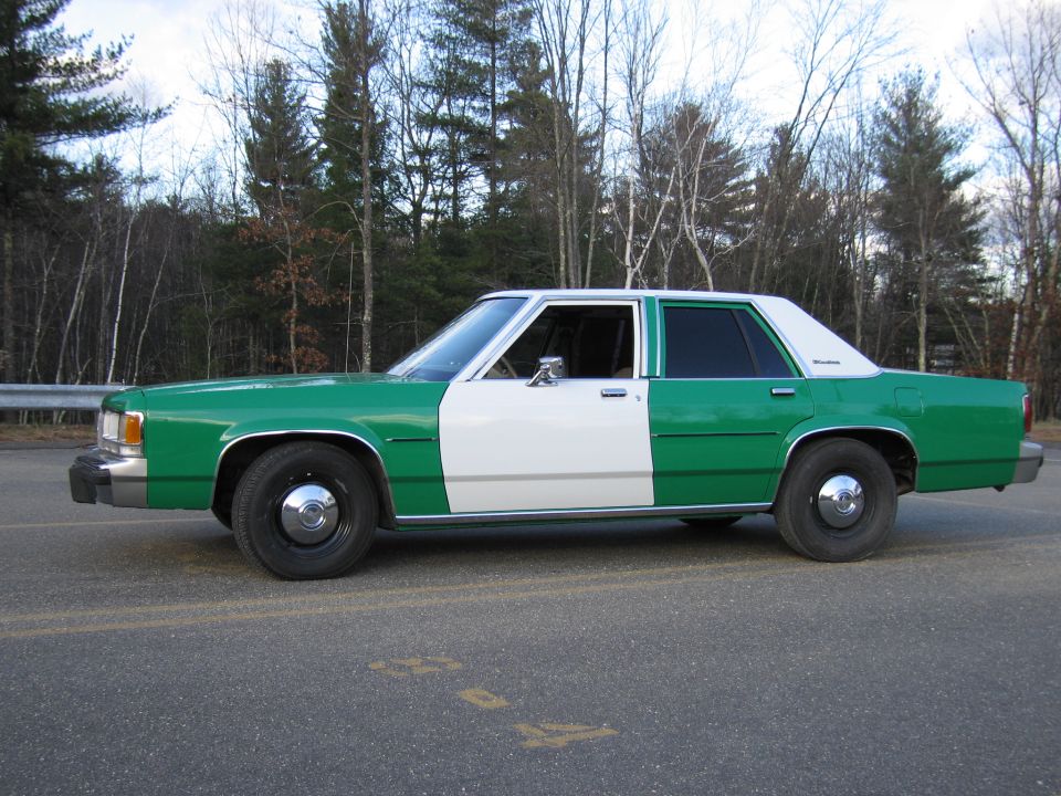 Green and white Ford sedan, front three-quarter, parked on lot by woods