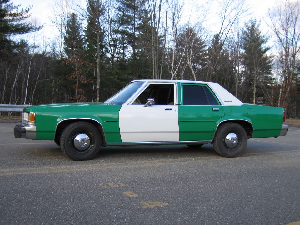 Green and white Ford sedan in profile parked on paved lot near trees