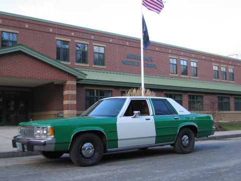 1991 Ford LTD Crown Victoria - Front three-quarter view of a green-and-white full-size Ford sedan parked outside a brick middle school with a flagpole.