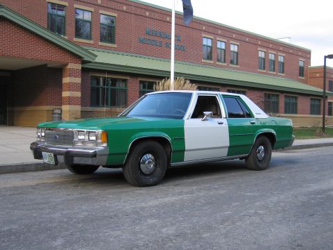 1991 Ford LTD Crown Victoria - Front three-quarter view of a green-and-white full-size Ford sedan parked at the curb in front of a brick middle school.