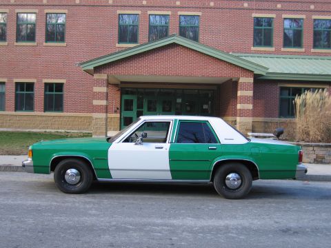 1991 Ford LTD Crown Victoria - Driver-side profile of a green-and-white full-size Ford sedan parked in front of a brick building with a green-roofed entrance.