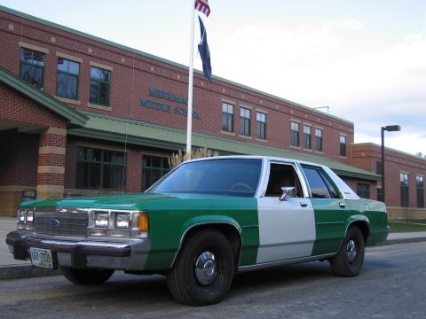 1991 Ford LTD Crown Victoria - Front three-quarter view of a green-and-white full-size Ford sedan with Ford grille emblem parked in front of a brick middle school.