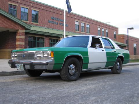 1991 Ford LTD Crown Victoria - green-and-white Ford LTD Crown Victoria four-door sedan sits at the curb outside a brick middle school building.