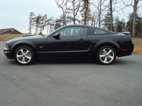 2007 Ford Mustang - Side profile of a black 2007 Ford Mustang GT coupe parked on an asphalt road with trees in the background.