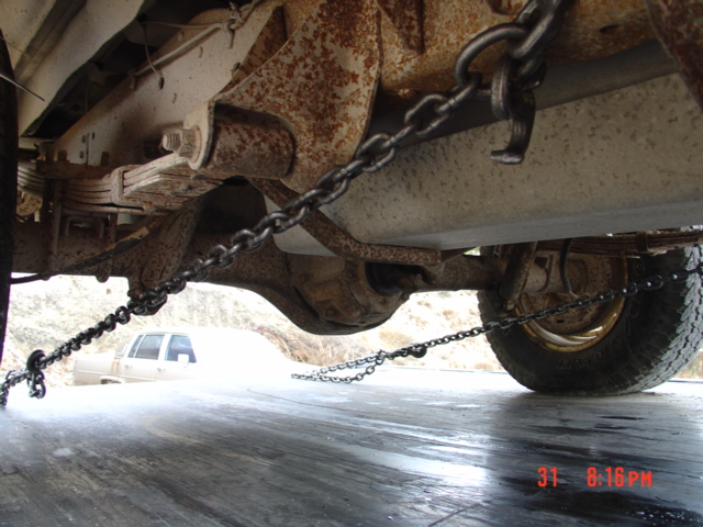 Undercarriage of a vehicle with chains attached to the frame and axle, another vehicle is visible in the background