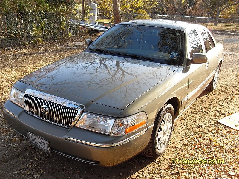 Front end of a gold Mercury Grand Marquis parked on a dirt lot. License plate is visible