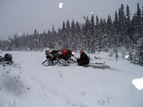 2000 Ford E350 - A person sits on a sled next to a snowmobile in a snow-covered forest with trees in the background.