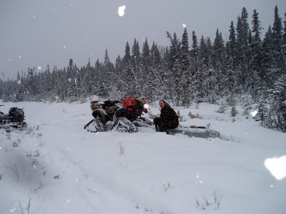 Two snowmobiles in a snowy forest with a person sitting on a sled