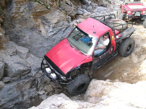1986 Toyota Hilux - A red pickup truck navigates a challenging rocky terrain with another off-road vehicle visible behind it.