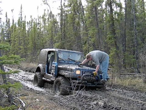 1991 Jeep Wrangler - A person works on a muddy blue 1991 Jeep Wrangler stuck on a forest trail.