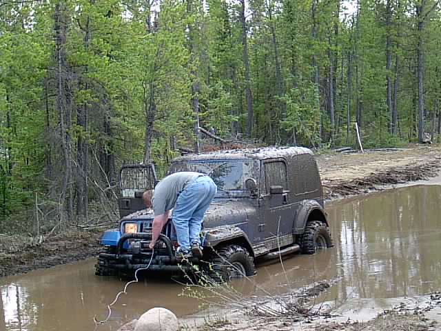 Mud-covered Jeep Wrangler stuck in a water hole while a person attaches a cable at the front bumper.
