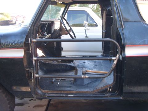 1978 Ford Bronco - Open doorway of a black Ford Bronco fitted with a tube half-door, showing steering wheel and carpeted interior.