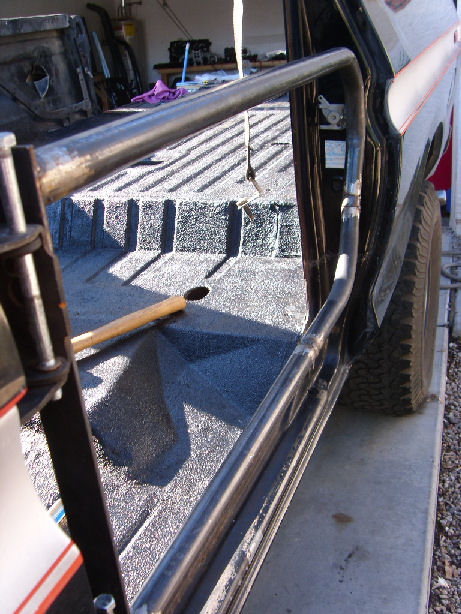 1978 Ford Bronco - A bare-metal welded tube door frame hung on a truck's door opening, seen from outside with sunlight showing the raw weld seams and hinge ta…