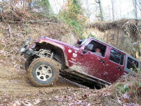 2007 Jeep Wrangler - A red Jeep Wrangler navigates a steep, muddy incline surrounded by trees and foliage.
