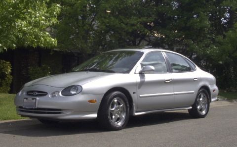 1998 Ford Taurus - A silver 1998 Ford Taurus is parked on a street with trees and foliage visible behind it.