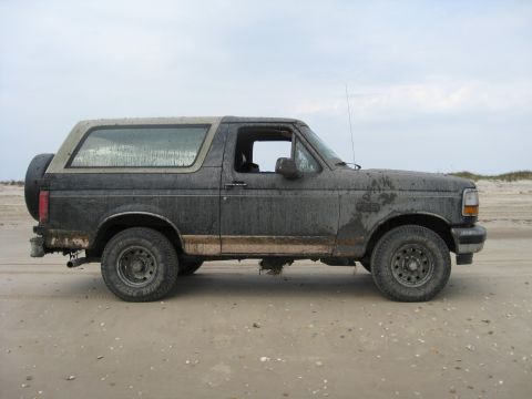 1994 Ford Bronco - A side view of a dirty Ford Bronco on a beach with a cloudy sky overhead.