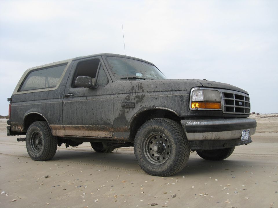 Mud-covered dark gray 1992 Ford Bronco on a sandy beach, viewed from the front three-quarter angle