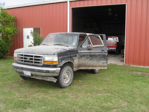1994 Ford Bronco - A muddy black Ford Bronco sits outside a red barn with its driver's door ajar, while another red truck is visible inside.