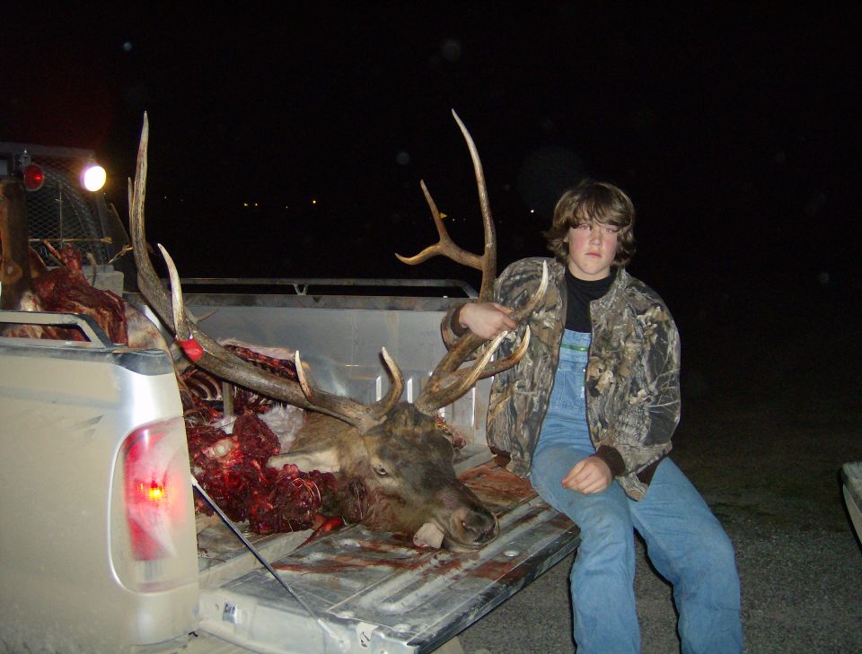 Kid in camo jacket and overalls sits on tailgate next to a large elk carcass in a truck bed. Another truck is partially visible to the left